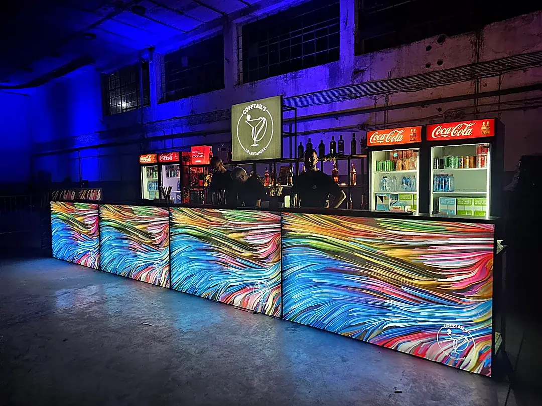 Bar de festival de mari dimensiuni, cu panouri multicolore și logo Cofftails iluminat, într-o hală industrială