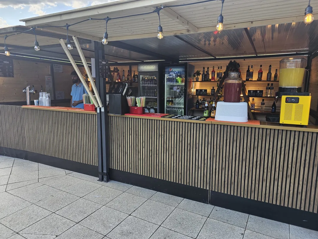 Bar mobil de exterior cu pergolă din lemn, dozatoare de limonadă și vitrine frigorifice, pregătit pentru o zi de eveniment