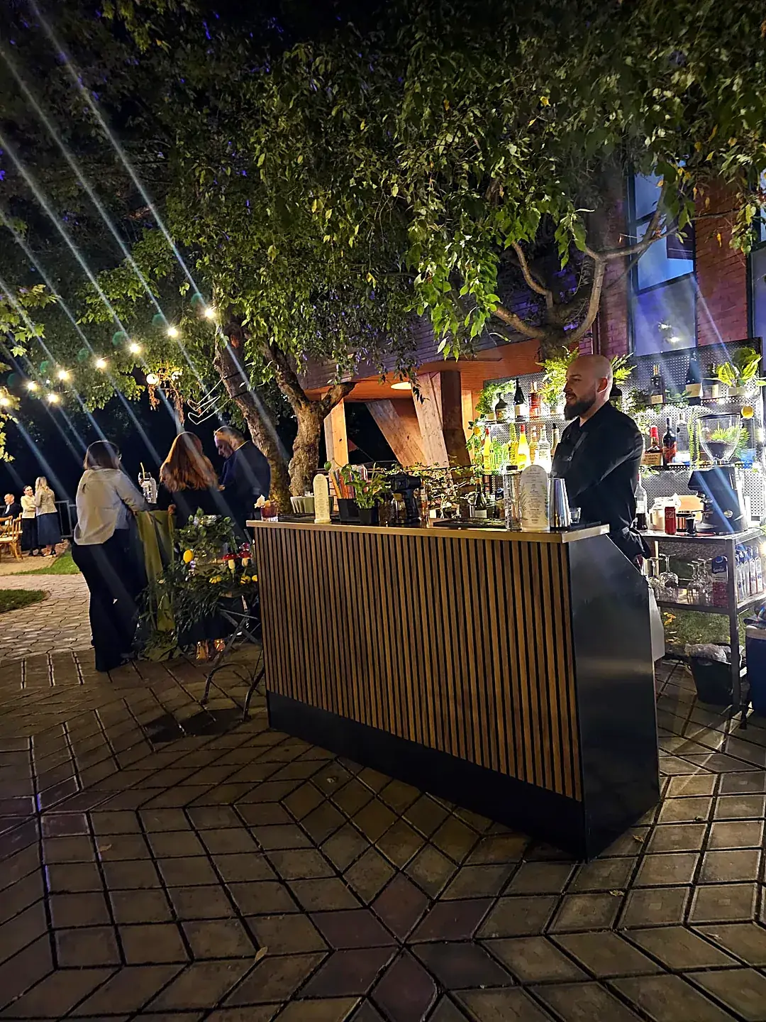 Bar mobil montat seara într-o grădină, cu barman, invitați și instalații de lumini, la un eveniment privat