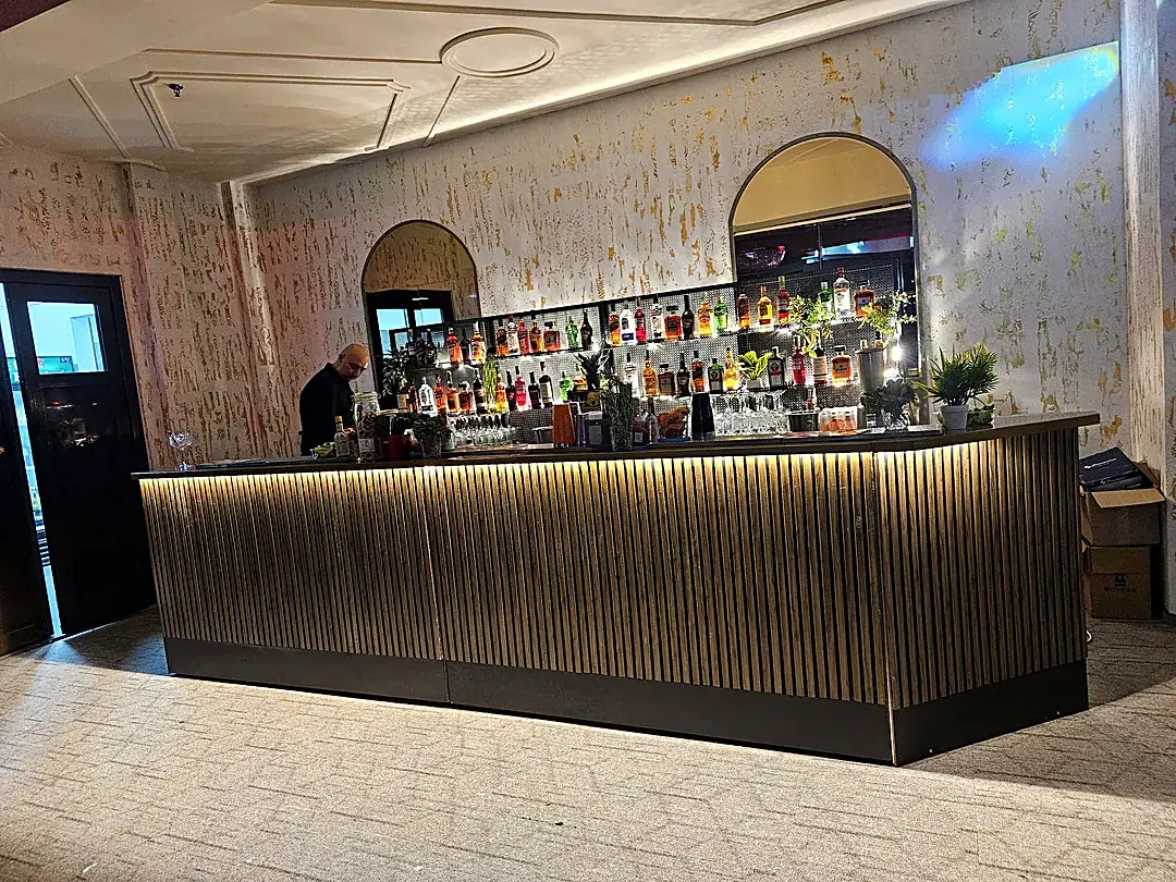 Bar mobil lung, în formă de L, cu riflaje din lemn și iluminare ambientală, montat într-o sală de evenimente